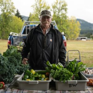 A farmer stands behind a table full of kale, peppers, and other fresh vegetables at the Port Alberni Farmers’ Market.