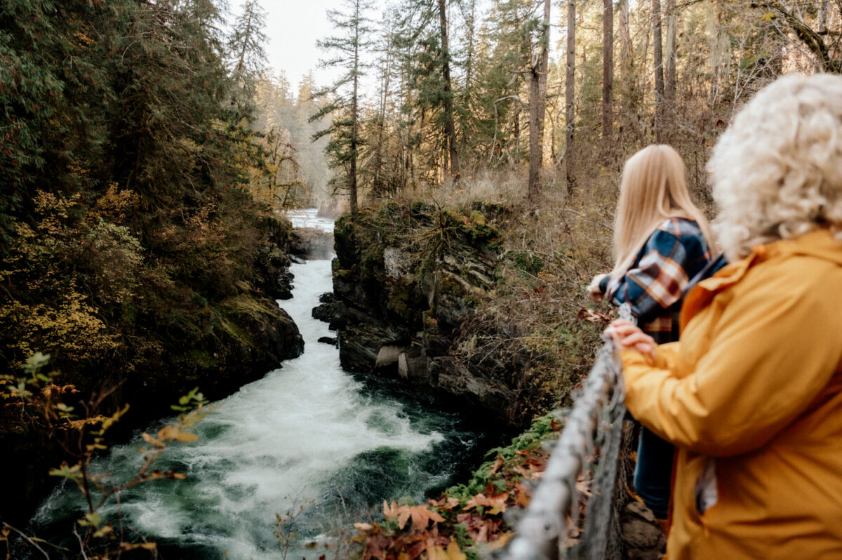 Two women looking out over the rushing water at Stamp Falls, surrounded by forest in early fall.
