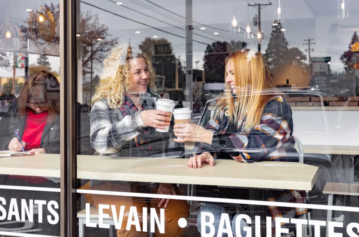 Two women smiling and cheersing coffee cups through a café window in downtown Port Alberni.