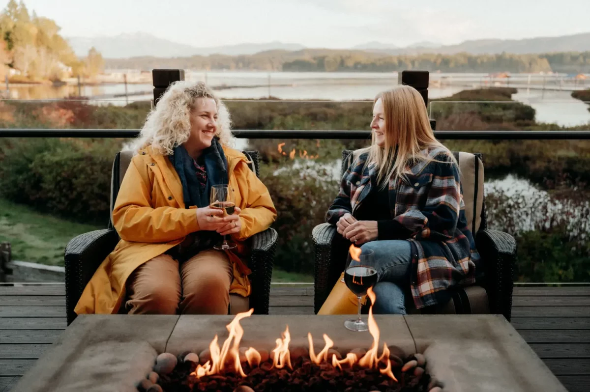 Two women in warm jackets sitting by an outdoor firepit with drinks, overlooking a calm inlet and fall landscape.