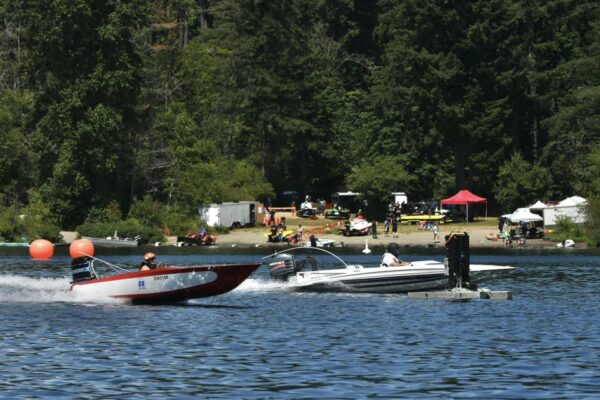 Crowd of families sitting on lakeside lawn chairs and blankets, watching boat races.