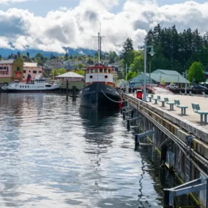 The boardwalk at Port Alberni Harbour Quay features green benches facing the water, with two docked ships nearby and colorful waterfront buildings, including the Maritime Centre, under a cloudy but bright sky.