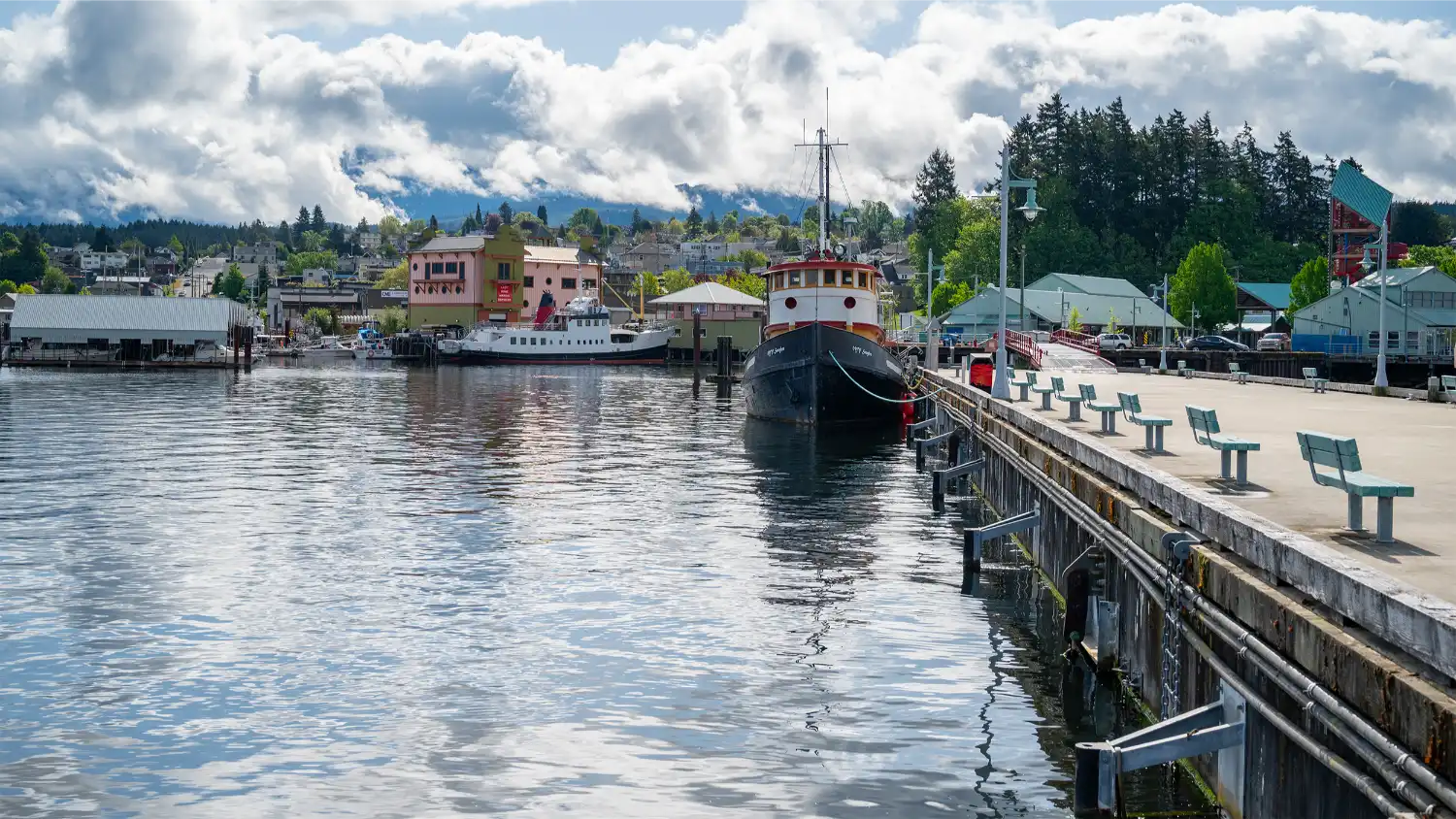The boardwalk at Port Alberni Harbour Quay features green benches facing the water, with two docked ships nearby and colorful waterfront buildings, including the Maritime Centre, under a cloudy but bright sky.