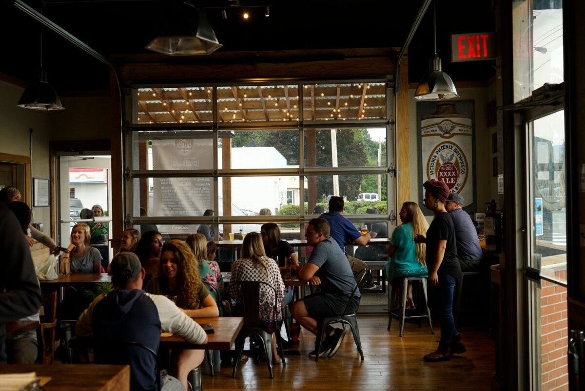 “People enjoying food and drinks inside Twin City Brewing, with warm lighting and a view of the street through the large garage-style window.”